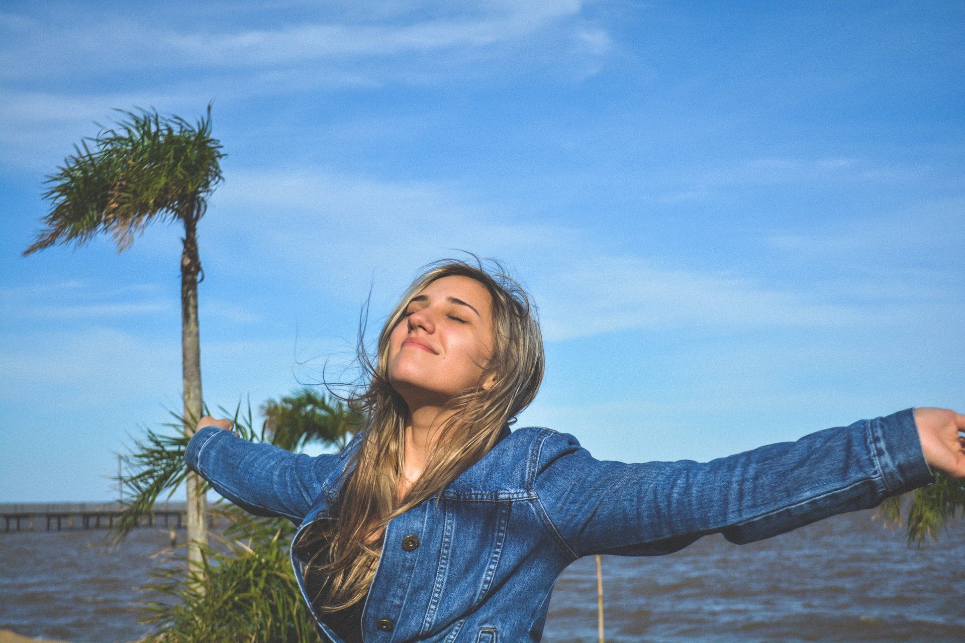 Woman with arms outstretched, eyes closed, smiling, wearing a denim jacket by a body of water under a blue sky.